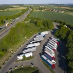 An aerial view of a truck rest area near a highway, surrounded by green fields and trees. Multiple semi-trucks with trailers are parked in an organized layout, with red, white, and blue truck cabs visible. The adjacent highway has vehicles traveling in both directions, with a few overpasses in the distance.