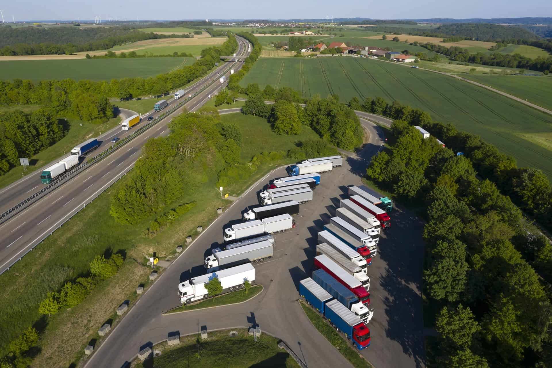 An aerial view of a truck rest area near a highway, surrounded by green fields and trees. Multiple semi-trucks with trailers are parked in an organized layout, with red, white, and blue truck cabs visible. The adjacent highway has vehicles traveling in both directions, with a few overpasses in the distance.