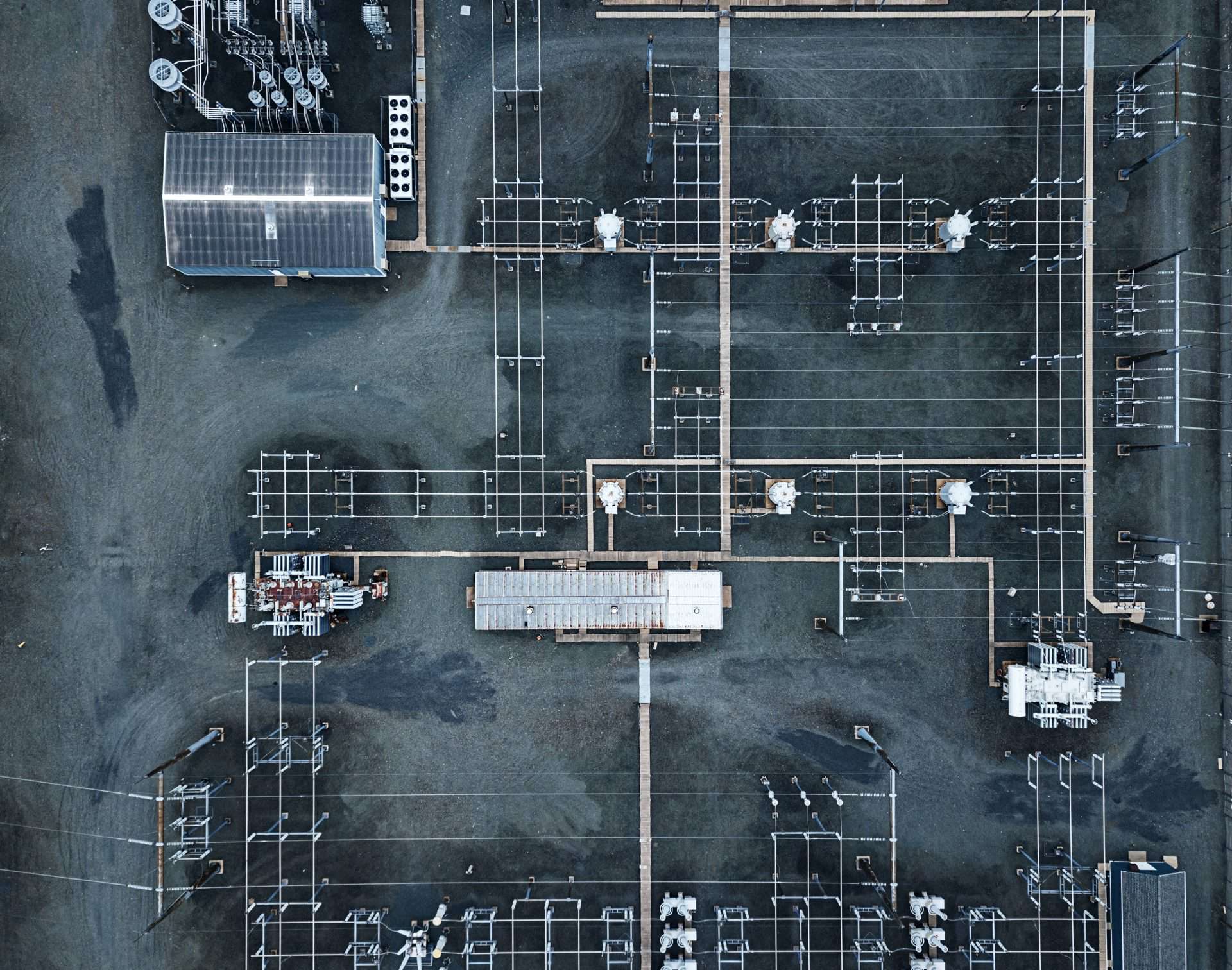 An aerial view of an electrical substation featuring a complex network of power lines, transformers, and metal frameworks arranged in a grid-like pattern. The site includes several large utility structures, such as a covered building and rows of equipment, spread across a gravel-covered area. The symmetrical arrangement of components and overhead wiring emphasizes the technical precision of the facility, showcasing its role in power distribution and infrastructure.