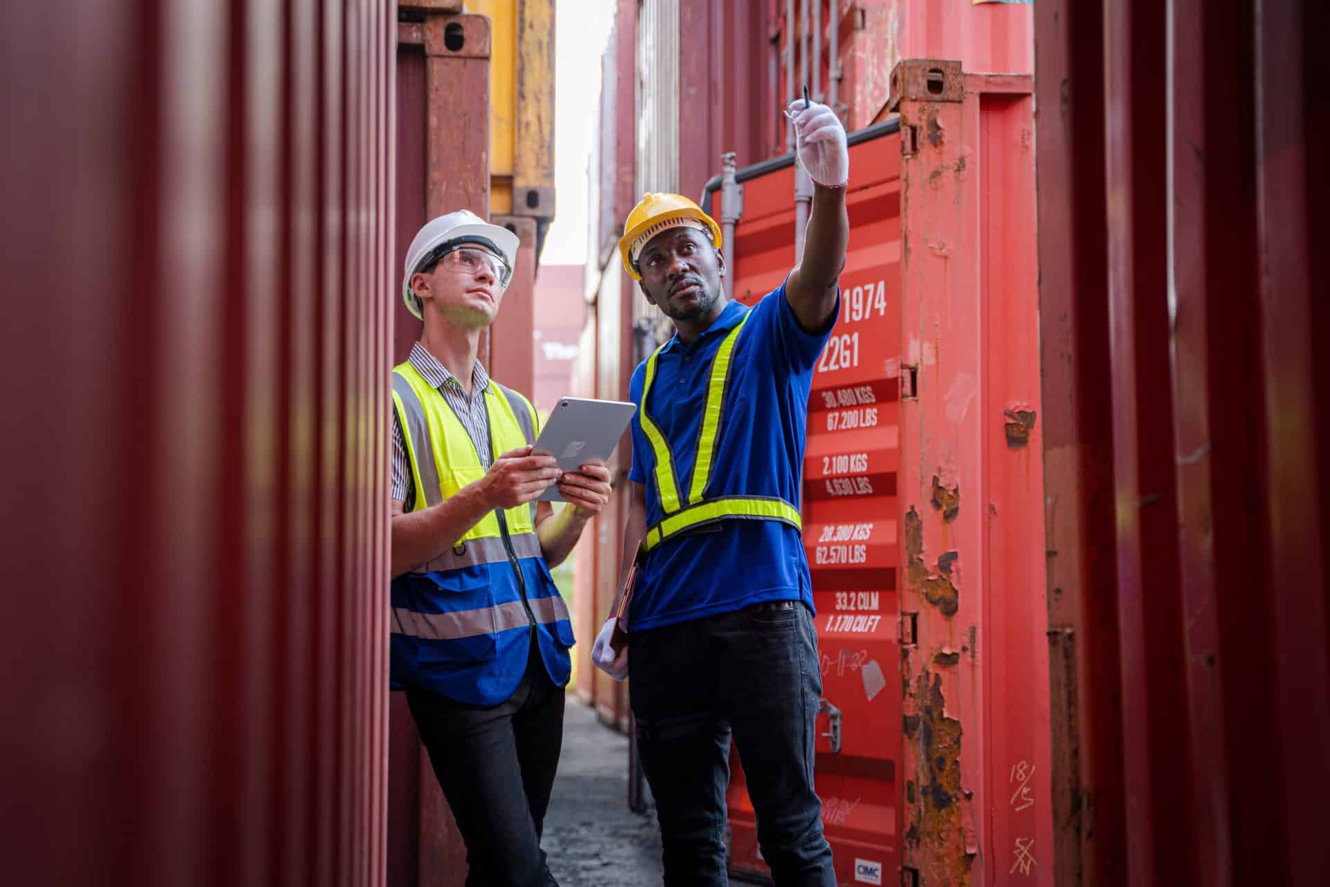 Two workers in a shipping yard stand between rows of red cargo containers, collaborating on a task. The worker on the left, wearing a white hard hat, safety glasses, and a high-visibility vest, holds a tablet and looks up attentively. The worker on the right, wearing a yellow hard hat, gloves, and a blue high-visibility vest, gestures toward a container, pointing something out.