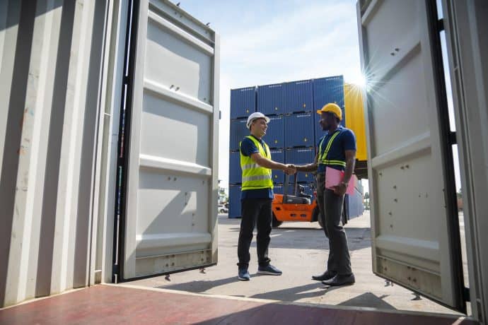 Two workers shake hands in a shipping yard, framed by the open doors of a cargo container. The worker on the left wears a white hard hat and a high-visibility vest, while the worker on the right wears a yellow hard hat, a reflective harness, and holds a clipboard. Behind them, stacks of blue and yellow shipping containers and an orange forklift are visible under bright sunlight.