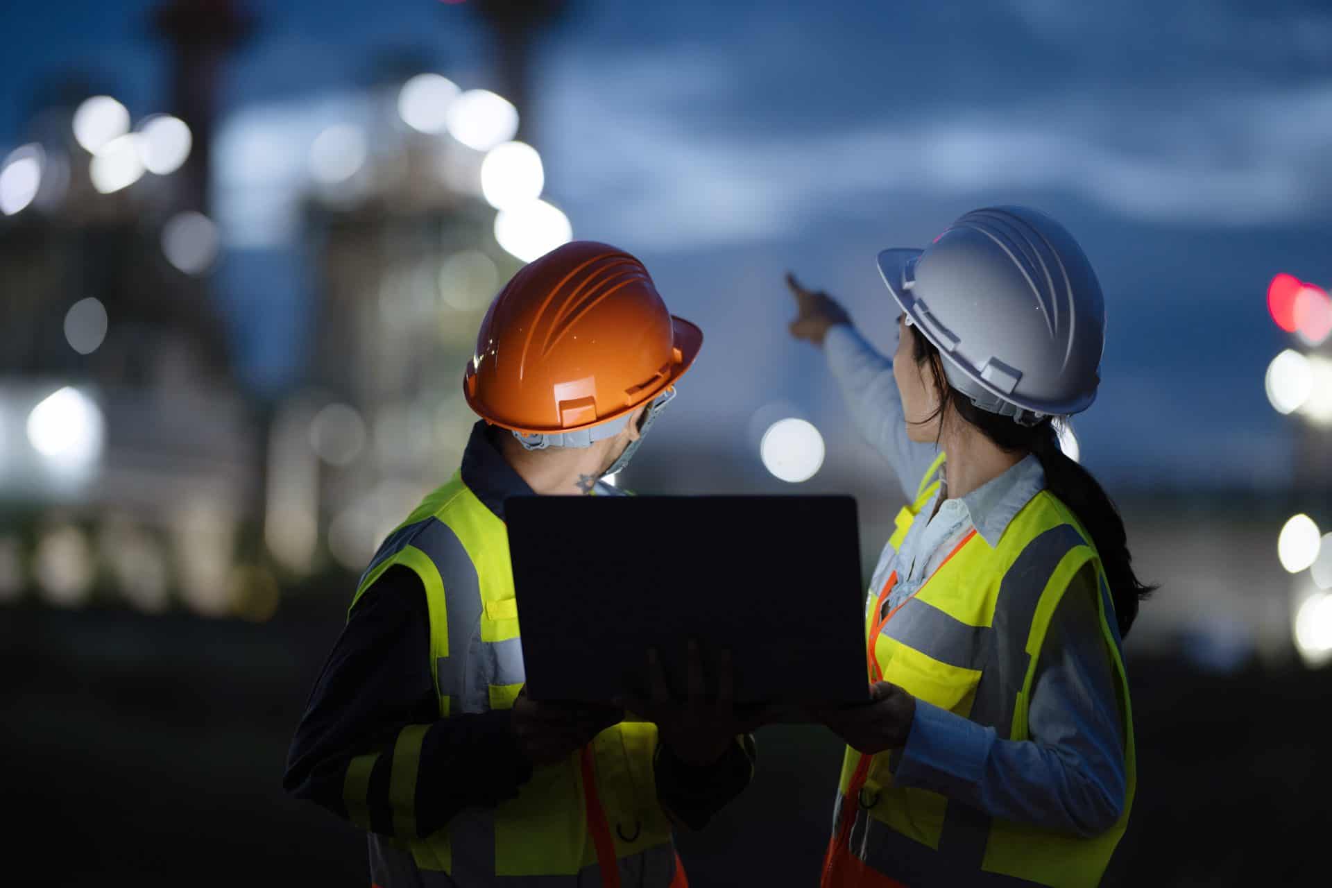 Two workers wearing high-visibility vests and hard hats stand outdoors at dusk, discussing or inspecting a site. One worker, wearing an orange helmet, holds a laptop, while the other, wearing a white helmet, points into the distance towards blurred industrial structures illuminated in the background.