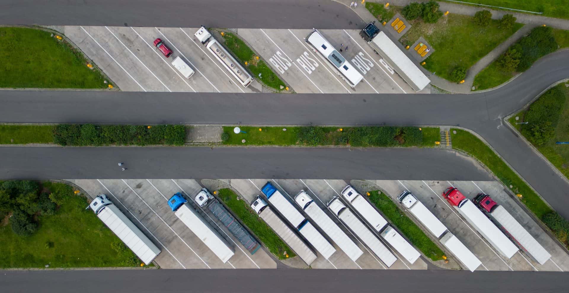 An aerial view of a roadside rest area featuring designated parking spaces for buses and trucks. On the upper half, several buses and a few smaller vehicles are parked in marked spaces labeled "BUS." In the lower half, trucks with trailers are parked in separate lanes, mostly aligned parallel to each other. The rest area includes green grass and shrubs separating the parking sections, with paved roads running through the area. A small picnic area with tables is visible in the top right corner, adding functionality to the rest stop.