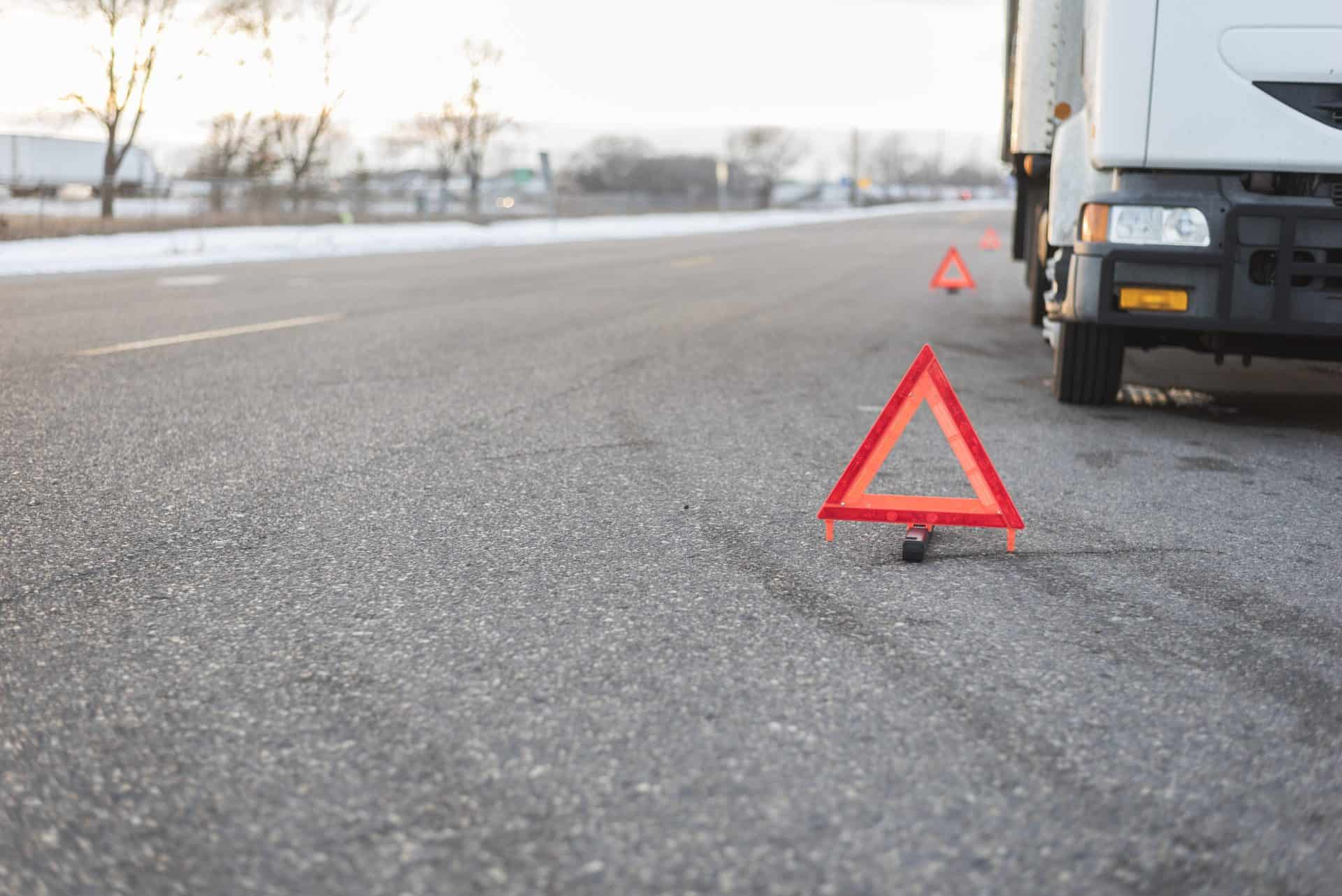 A roadside safety scene showing a truck pulled over on a wide road with red triangular warning signs placed at intervals behind it. The reflective triangles are positioned to alert other drivers of the stationary vehicle. The road stretches into the distance, with a snowy landscape and leafless trees visible in the background under soft daylight.