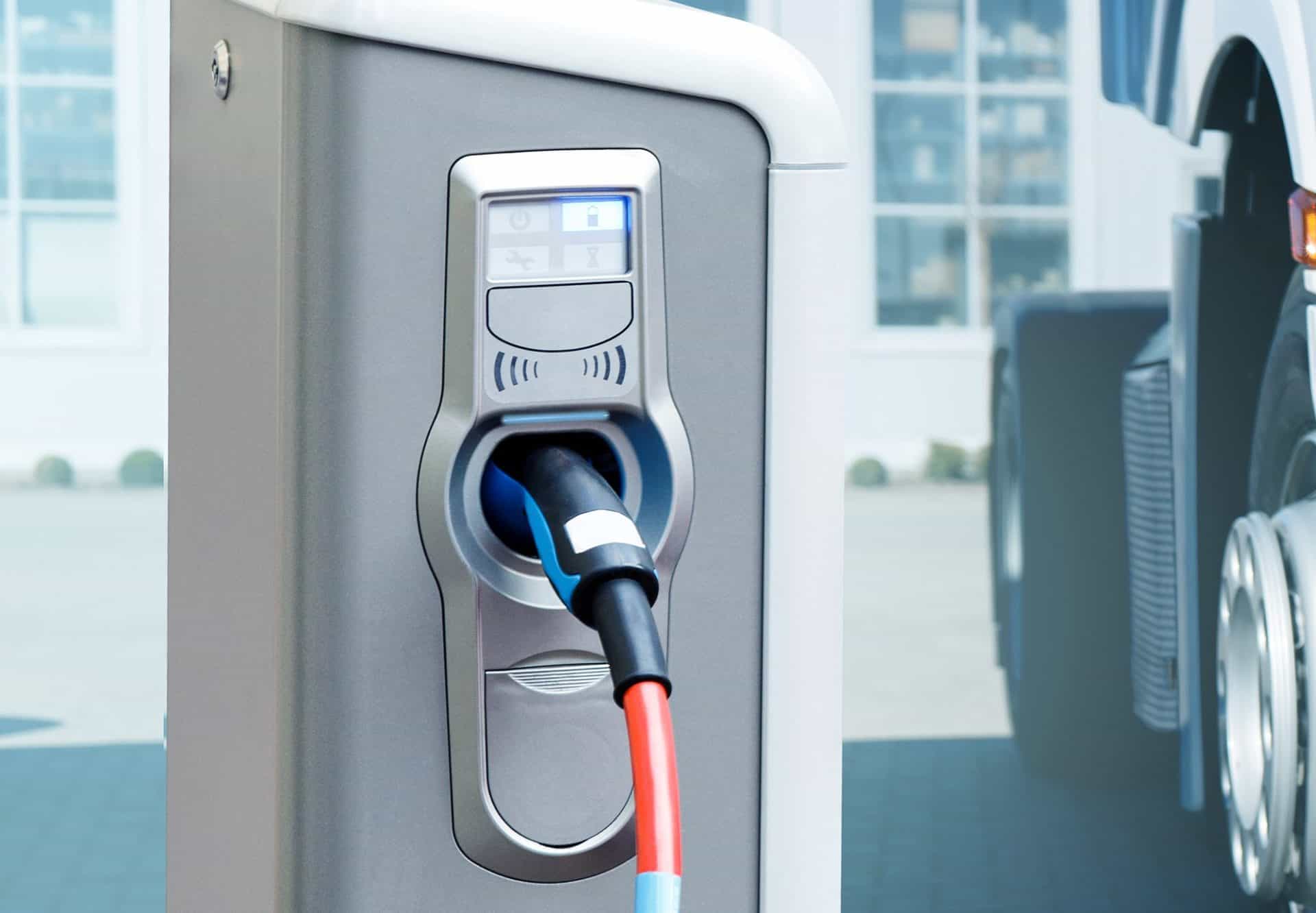 A close-up view of an electric vehicle charging station with a charging cable plugged into the port. The charger has a sleek metallic design, with icons and indicators above the port displaying charging status. In the background, part of a large vehicle, likely a truck, is visible, suggesting a focus on electric commercial vehicles.