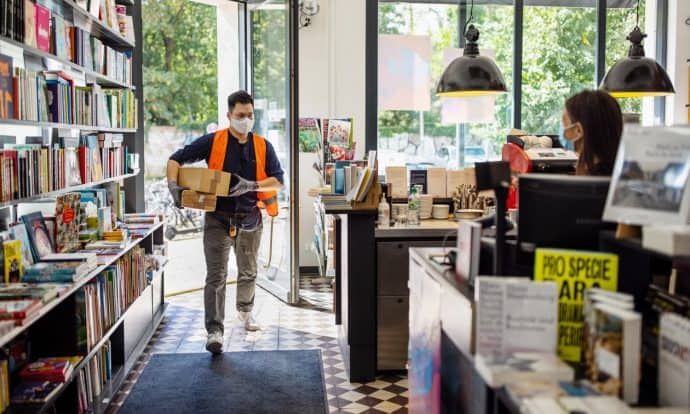 A delivery person wearing a high-visibility orange vest, mask, and gloves carries a package into a brightly lit store filled with books and stationery. Another individual behind the counter, also wearing a mask, looks on as the delivery is made.